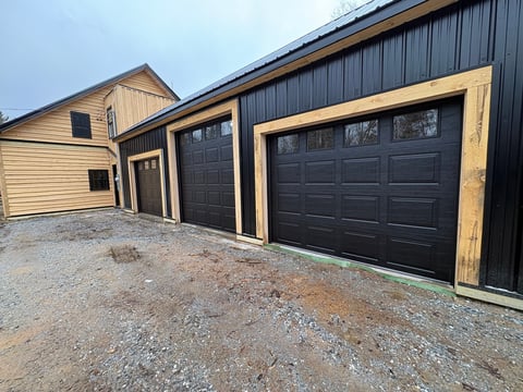 Modern black garage with wood trim and gravel driveway, attached to cream-colored house with dark roof
