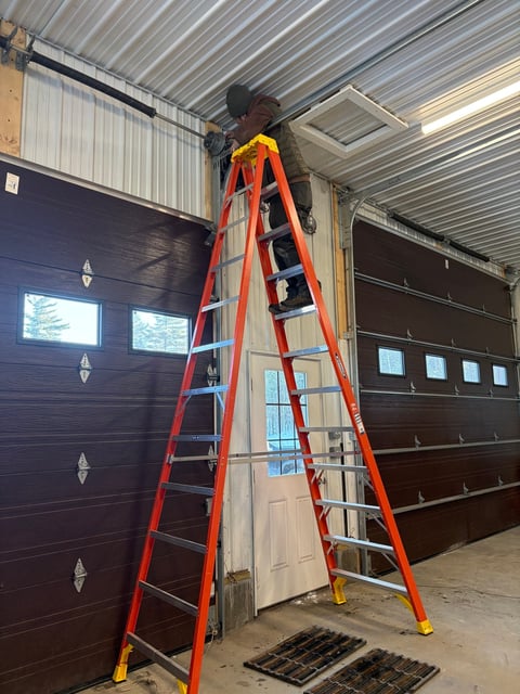 Worker on tall red ladder performing maintenance on garage door in industrial warehouse setting