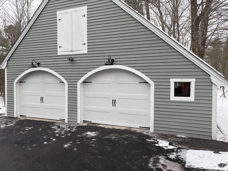 Gray detached garage with two white arched garage doors and bare winter trees in background