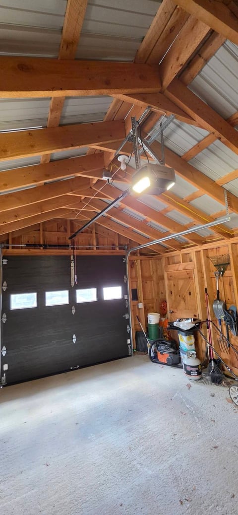 Interior of a wooden garage with exposed beams, black garage door with windows, concrete floor, LED work light, and storage items on the right side