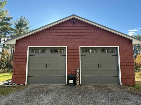 Two-car garage with two green garage doors, small windows, and trees surrounding it on a clear day