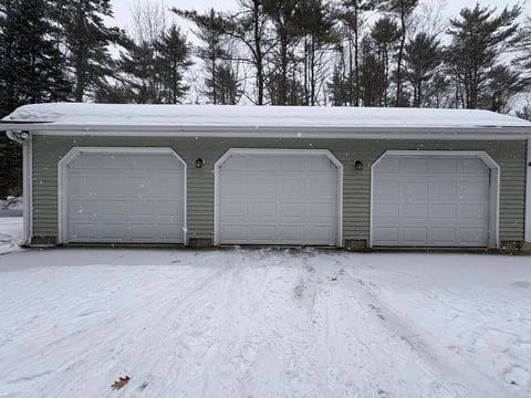 A three-car residential garage with white doors attached to a light-colored house, surrounded by evergreen trees with snow on the ground