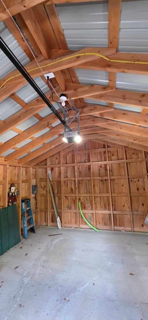 Interior of unfinished garage under construction with wooden framing, metal roof panels, concrete floor, and work equipment