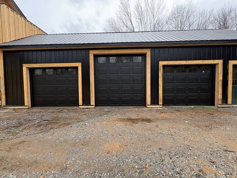 Three black garage doors with natural wood trim on a metal building with gravel driveway