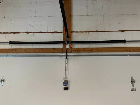 Garage door opener motor mounted on wooden beam with rails and cable system attached to white garage door panels