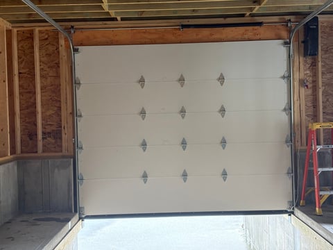 White overhead garage door with metal hardware mounted in an open garage frame with brick walls and wooden ceiling beams