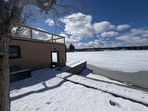 Modern wooden cabin on snowy beach beside frozen lake under blue sky with white clouds