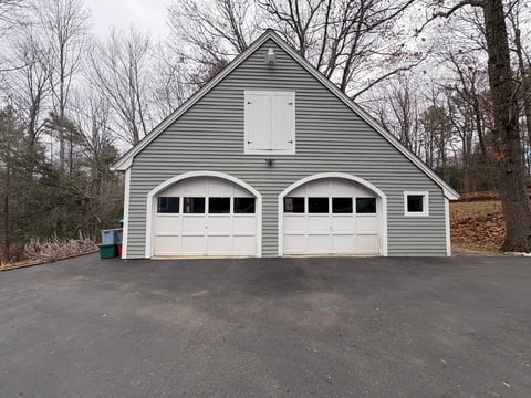 Two-car garage with white doors and gray siding surrounded by bare trees and driveway