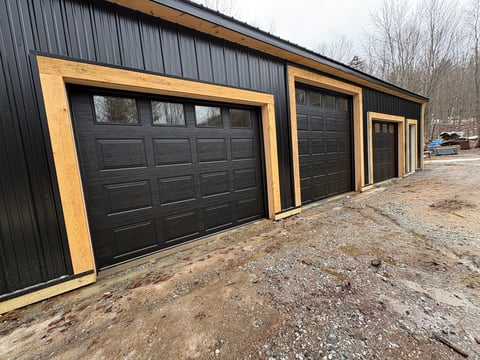 Modern three-bay garage with black steel panels and wooden trim doors on gravel driveway