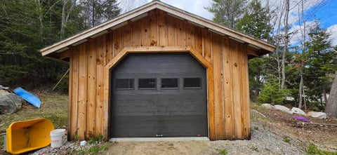 New wooden garage with dark gray garage door, surrounded by trees and landscaping on a residential property