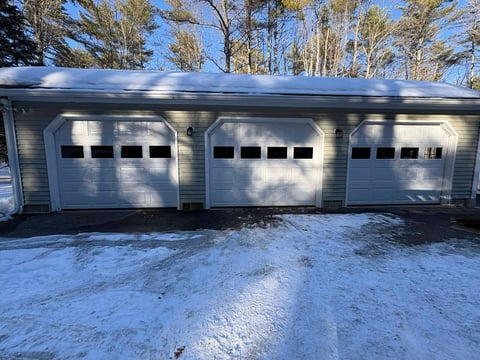 Three white garage doors on a residential building with bare trees and snow on the ground